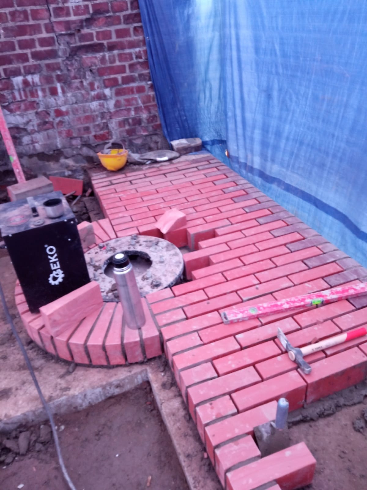 Układanie czerwonej cegły klinkierowej wokół okrągłego elementu, widoczne narzędzia murarskie, poziomica, tło z ceglanej ściany i niebieskiej plandeki.