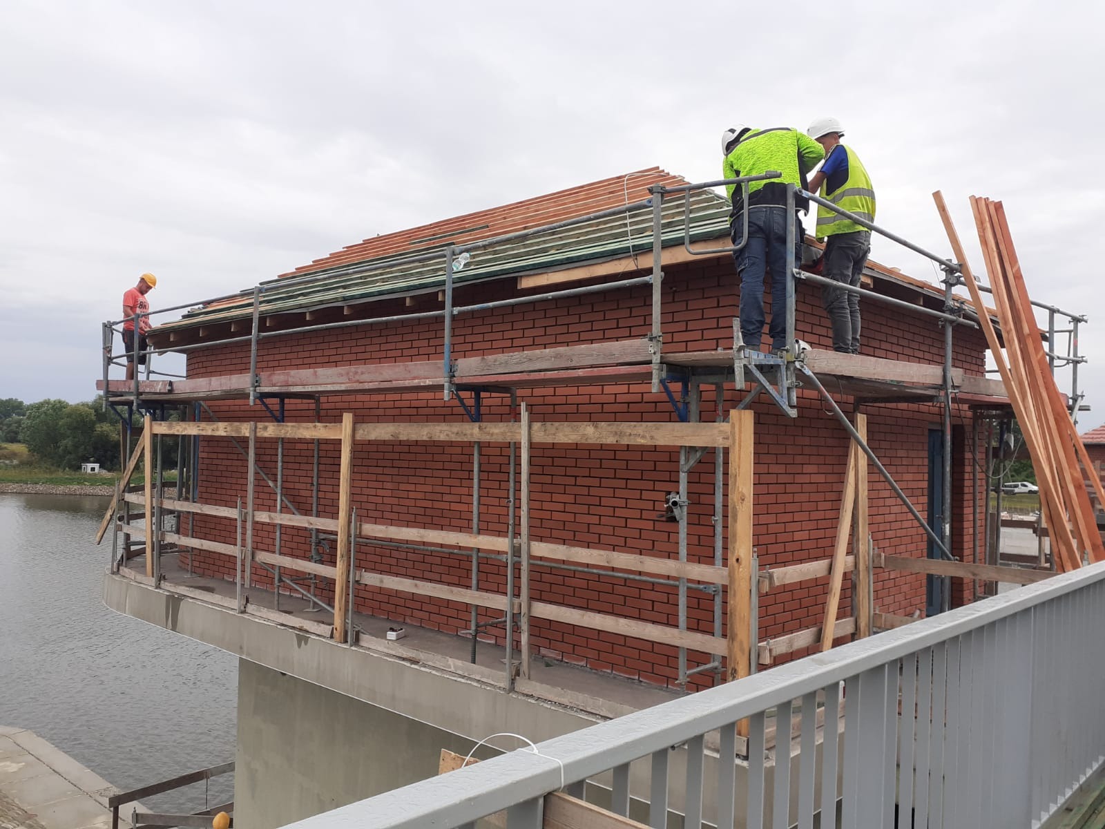 Budowa domu z czerwonej cegły na platformie nad wodą, robotnicy na rusztowaniu układają dachówkę.