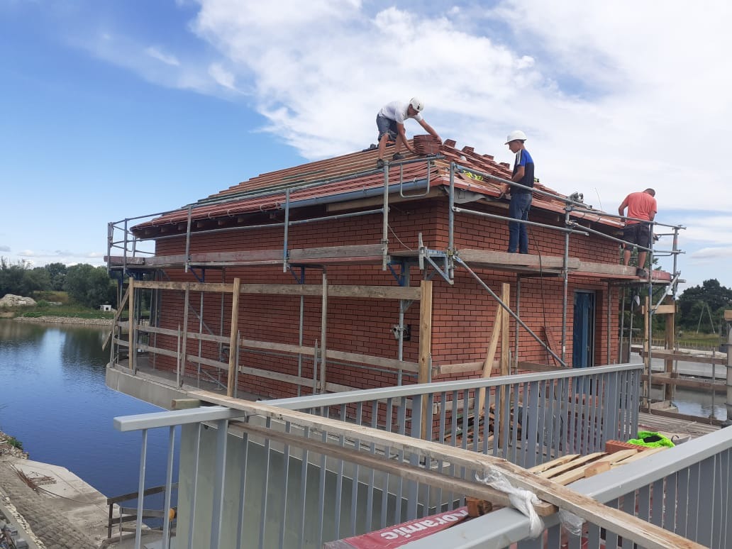 Murarze układają czerwoną dachówkę na dwukondygnacyjnym budynku z czerwonej cegły, otoczonego rusztowaniami, z widokiem na jezioro i błękitne niebo.