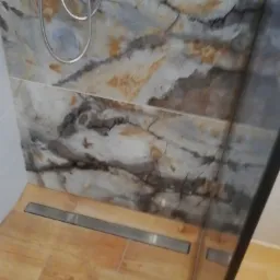 Wykończona kabina prysznicowa z odpływem liniowym, marmurowymi płytkami ściennymi i drewnopodobną podłogą z widocznym chromowanym zestawem natryskowym.