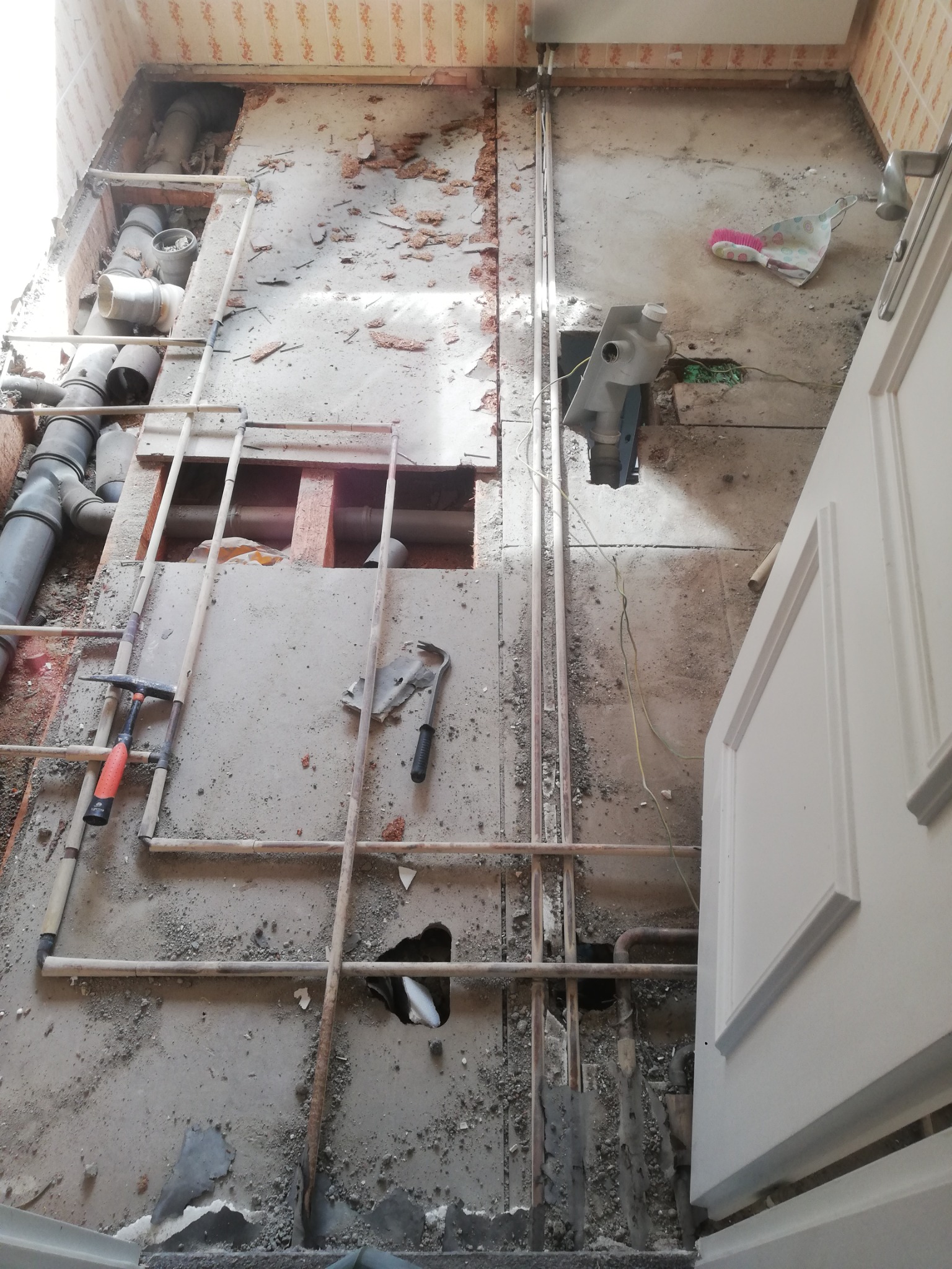 Podłoga w trakcie demontażu z widocznymi rurami, przewodami elektrycznymi, narzędziami i fragmentem drzwi. Widoczne ślady po starych płytkach i usuniętej warstwie podłogi.