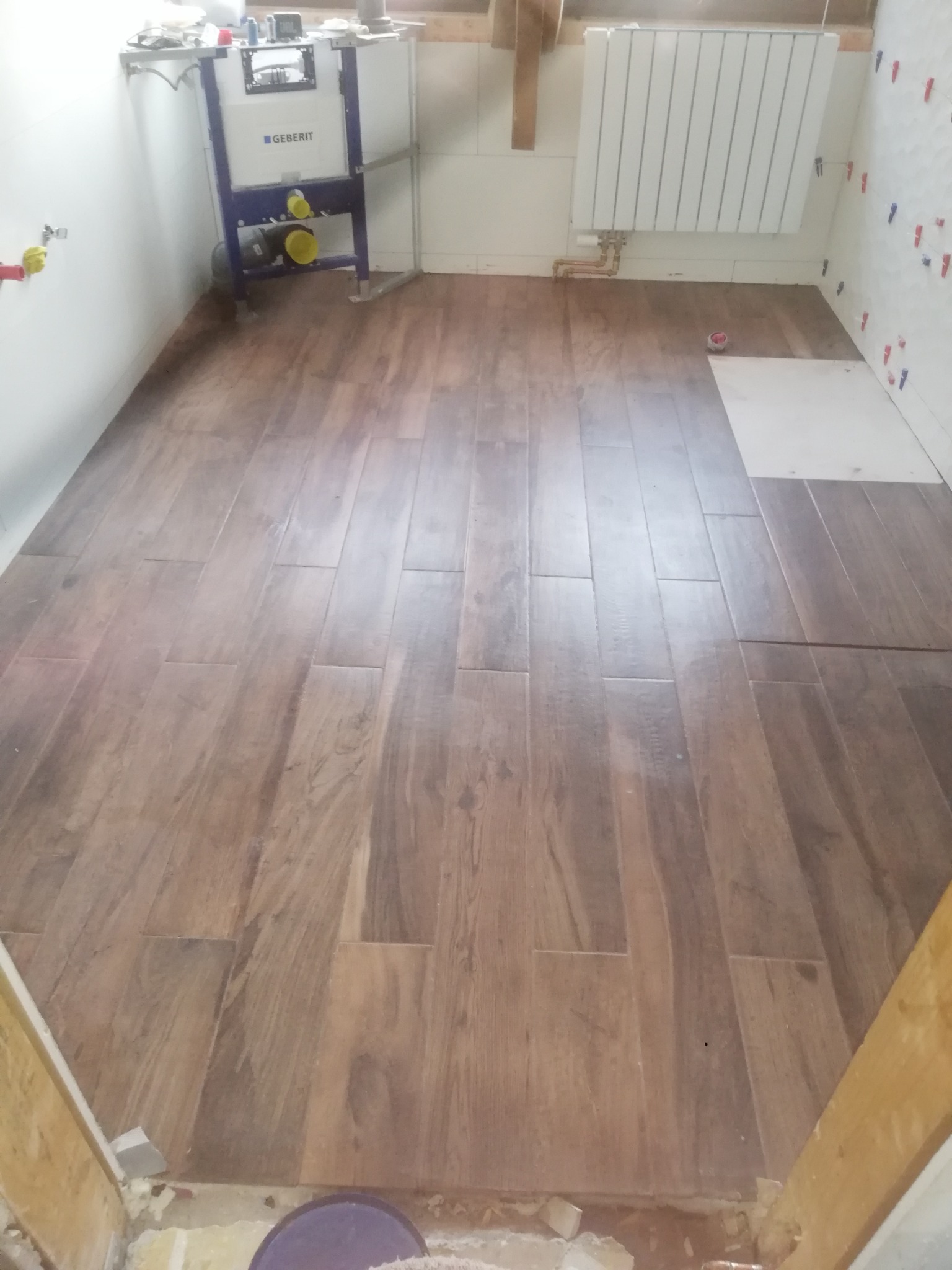 Ułożona podłoga z płytek imitujących drewno w trakcie remontu łazienki, widoczny stelaż podtynkowy WC Geberit, grzejnik na ścianie i krzyżyki dystansowe do płytek na ścianie.