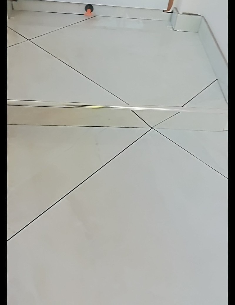 Białe, błyszczące płytki podłogowe ułożone pod kątem, z widoczną chromowaną listwą przypodłogową i elementem dekoracyjnym w rogu.