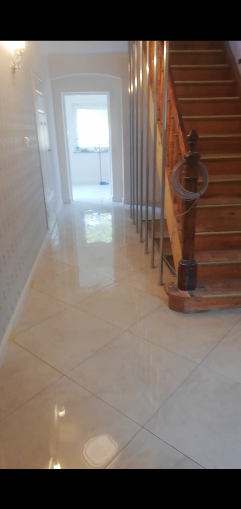 Korytarz z nowo położonymi, błyszczącymi, kremowymi płytkami podłogowymi ułożonymi w karo, widoczne schody z drewnianą balustradą i metalowymi prętami, ściany z delikatnym wzorem, drzwi wejściowe...