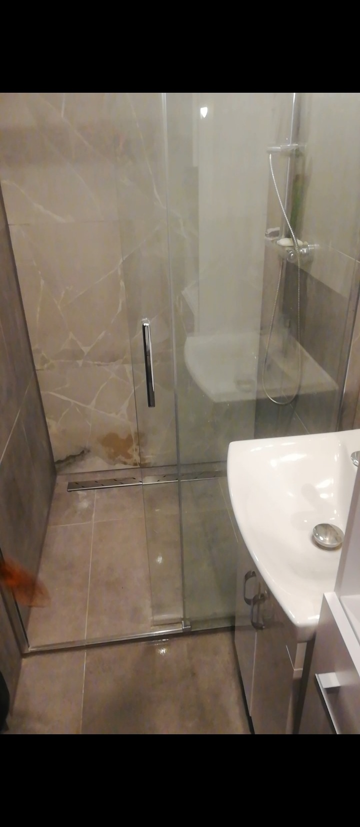 Nowoczesna kabina prysznicowa z szklanymi drzwiami i chromowanym uchwytem, widoczna umywalka z szafką, płytki imitujące marmur na ścianach, brązowe płytki podłogowe, częściowo widoczna dłoń w lewym...