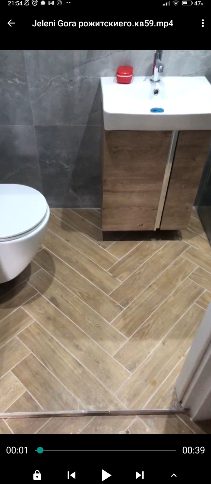 Ukończona łazienka z drewnopodobnymi płytkami ułożonymi w jodełkę na podłodze, szare płytki na ścianie, biała umywalka z szafką z drewnianym frontem oraz toaleta.