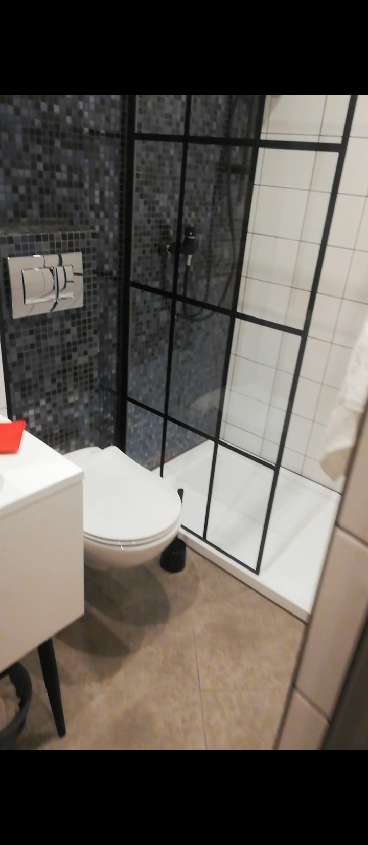 Nowoczesna łazienka z kabiną prysznicową typu walk-in z czarnymi profilami, mozaiką na ścianie i podwieszaną toaletą.