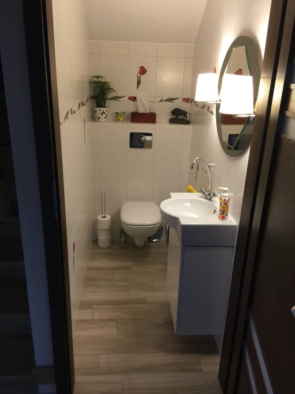 Niewielka łazienka z białymi płytkami zdobionymi motywem tulipanów, drewnopodobne płytki na podłodze, biała umywalka z szafką, okrągłe lustro z oświetleniem, widok zza otwartych drzwi.