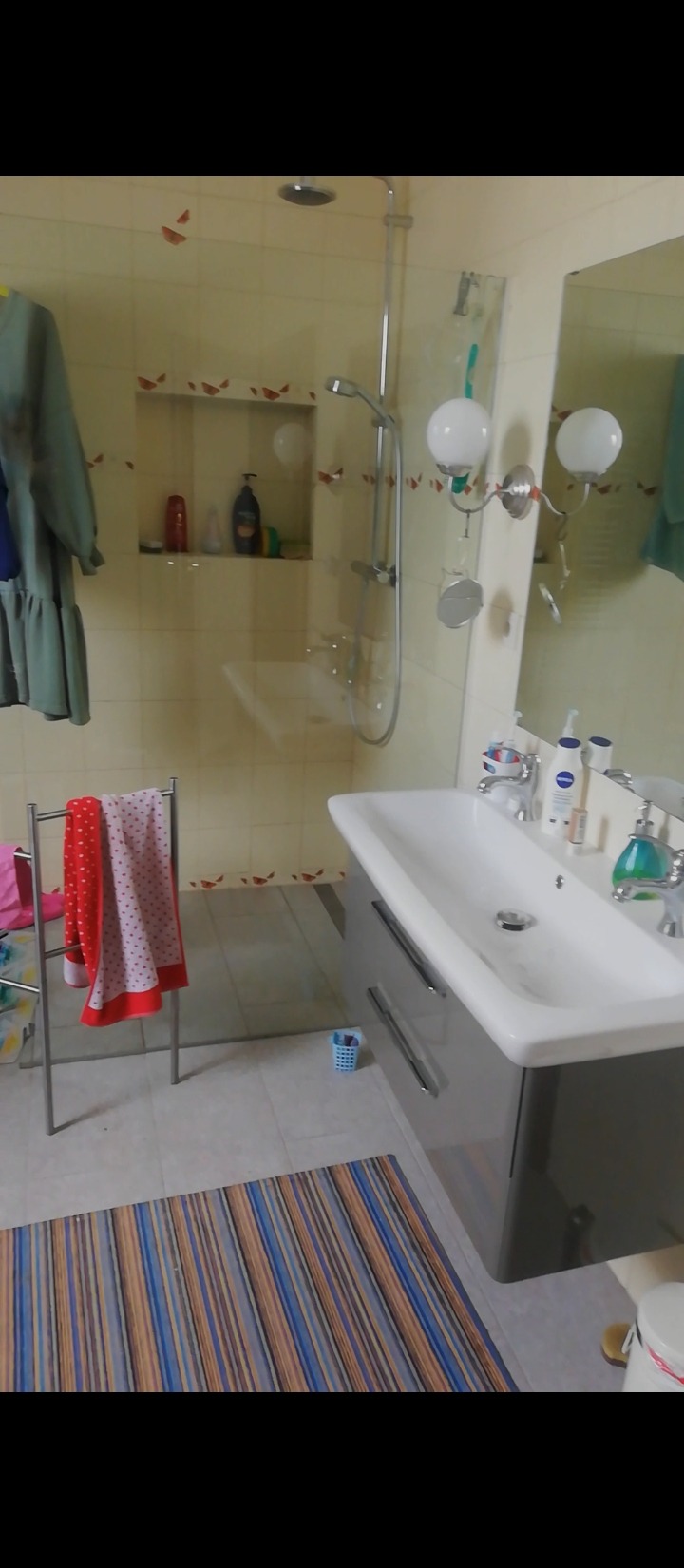 Umywalka z szarą szafką, baterią i kosmetykami w łazience z kabiną prysznicową, ręcznikiem na wieszaku i wzorzystym dywanikiem na podłodze.