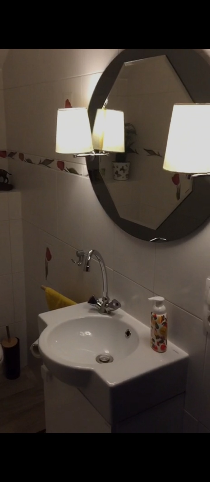 Umywalka narożna z białą szafką, chromowaną baterią i dozownikiem mydła w łazience z białymi płytkami i ozdobnym paskiem z motywem kwiatowym. W tle okrągłe lustro z oświetleniem.