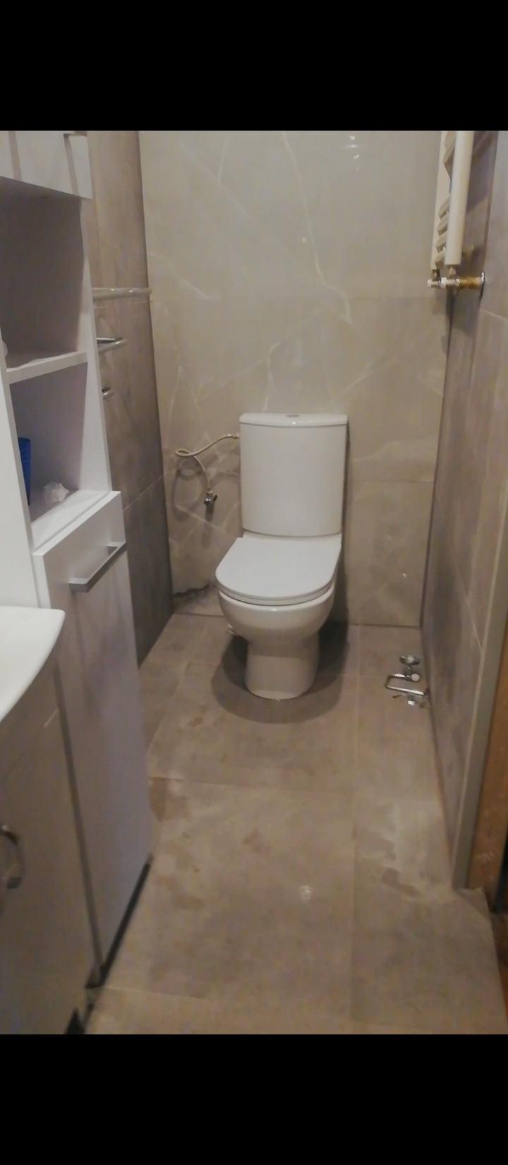 Wykończona łazienka z beżowymi płytkami imitującymi marmur na ścianach i podłodze, biała toaleta kompaktowa, biała szafka pod umywalkę z otwartymi półkami i chromowanymi relingami na ręczniki.