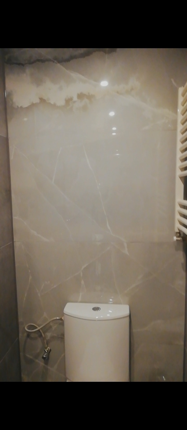 Wykończona łazienka z toaletą i grzejnikiem drabinkowym, ściany wyłożone płytkami imitującymi marmur w odcieniach beżu i szarości.