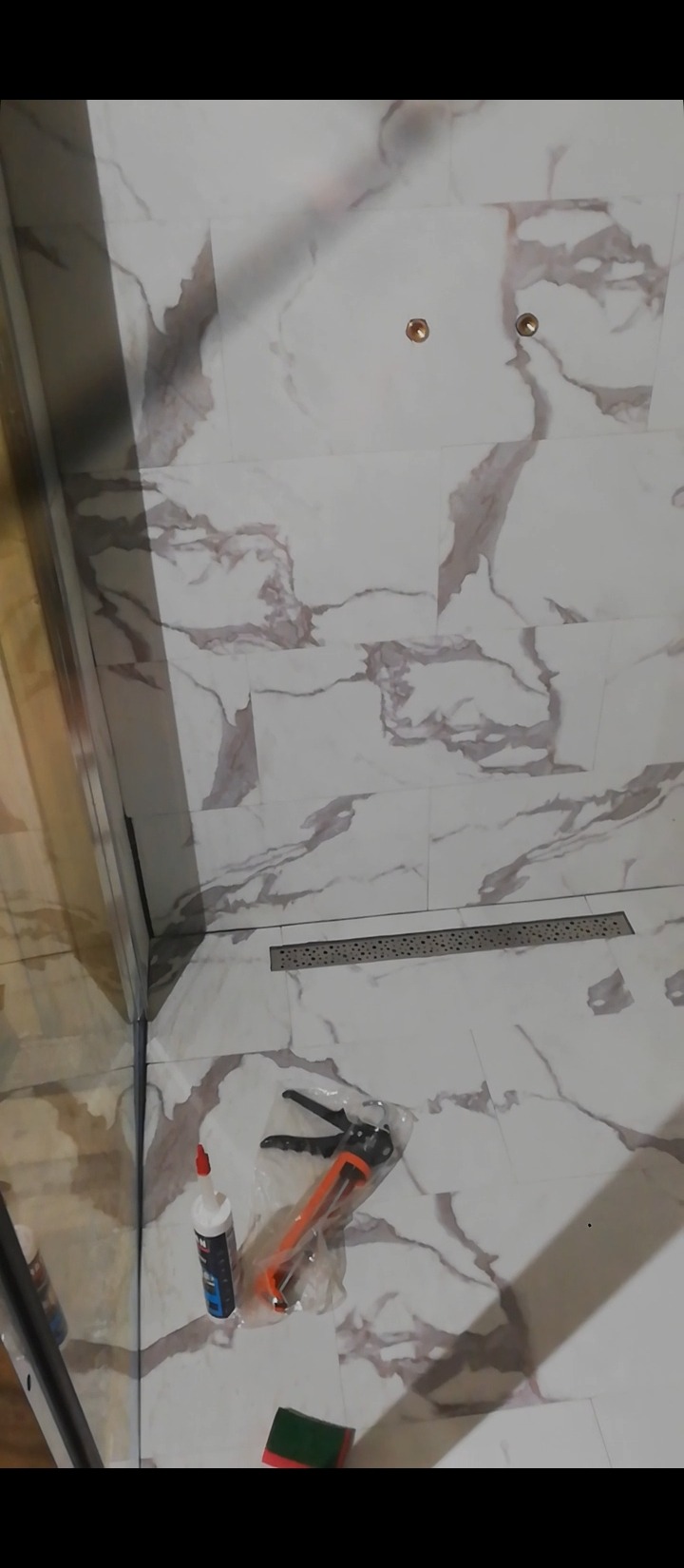 Fragment łazienki w trakcie remontu, widoczna kabina prysznicowa, odpływ liniowy w podłodze, marmurowe płytki na ścianach, narzędzia glazurnicze i chemia budowlana przygotowane do uszczelniania...