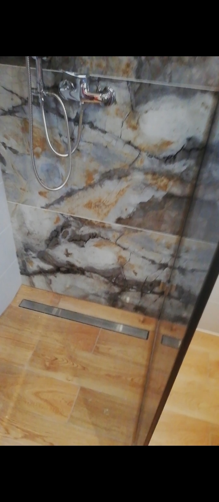 Wykończona kabina prysznicowa z odpływem liniowym, marmurowymi płytkami ściennymi i drewnopodobną podłogą z widocznym chromowanym zestawem natryskowym.