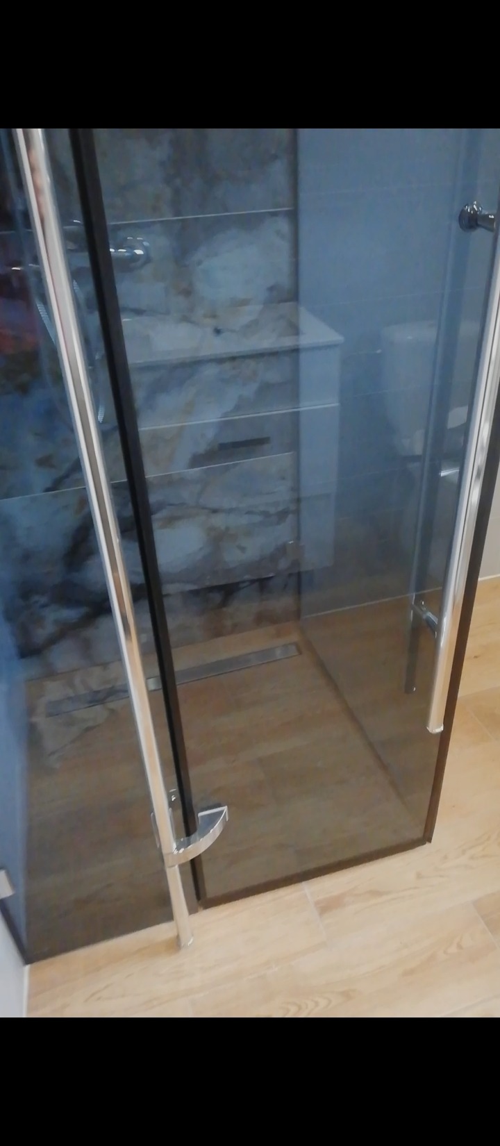 Kabina prysznicowa z czarnymi profilami i chromowanymi uchwytami, widoczny odpływ liniowy w posadzce, marmurowe płytki na ścianach wewnątrz kabiny, drewnopodobne płytki podłogowe.