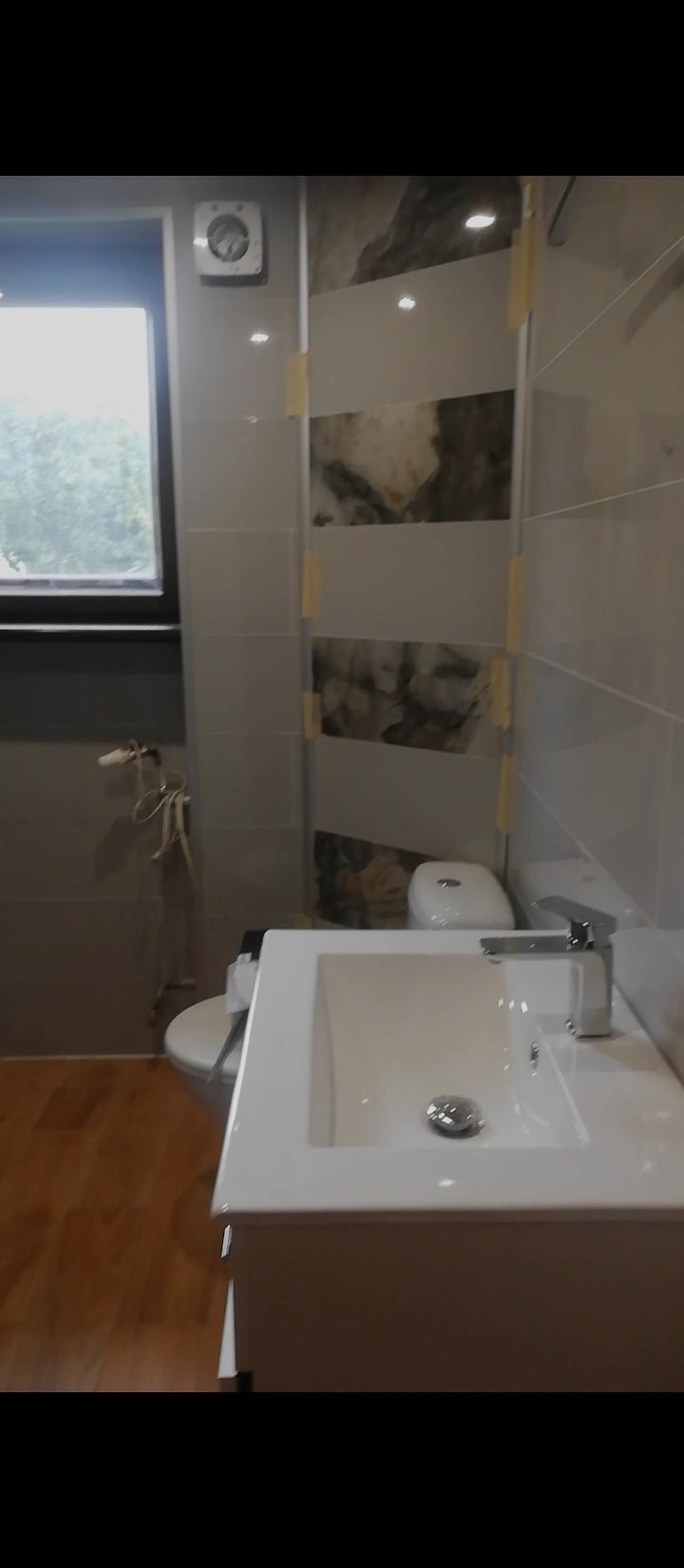 Nowoczesna łazienka w trakcie wykańczania: biała umywalka z chromowaną baterią, częściowo ułożone płytki ścienne z motywem marmuru zabezpieczone taśmą, drewnopodobna podłoga i okno z widokiem...