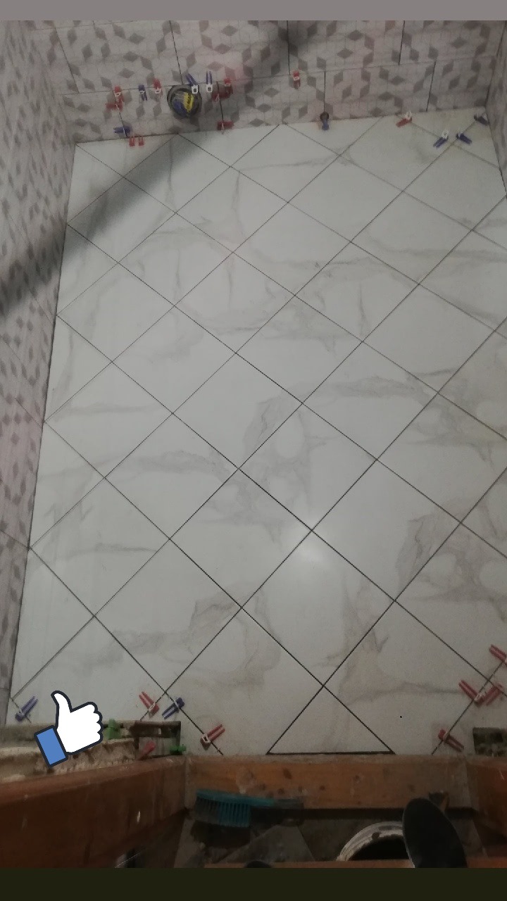 Układanie białych płytek podłogowych w łazience pod kątem 45 stopni, z użyciem klipsów poziomujących i krzyżyków dystansowych, widoczne narzędzia i fragment brodzika.