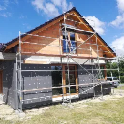 Budowa domu jednorodzinnego: ocieplenie ścian styropianem grafitowym, murowanie ścian z cegły ceramicznej, rusztowania na zewnątrz, błękitne niebo w tle.