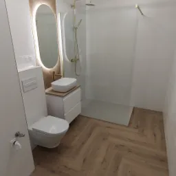 Nowoczesna łazienka z drewnianą podłogą ułożoną w jodełkę, owalnym lustrem z podświetleniem LED, białą umywalką nablatową i kabiną prysznicową ze złotą armaturą, oświetlona okrągłą lampą sufitową.