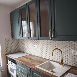 KONKRET HOME SPÓŁKA Z OGRANICZONĄ ODPOWIEDZIALNOŚCIĄ - Kuchnia po remoncie: zielone szafki z frezowanymi frontami i złotymi uchwytami, mozaika z białych płytek ze złotą fugą nad blatem z drewnopodobnego laminatu.