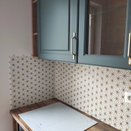 KONKRET HOME SPÓŁKA Z OGRANICZONĄ ODPOWIEDZIALNOŚCIĄ - Kuchnia po remoncie: mozaika z geometrycznym wzorem w odcieniach złota i bieli, złote uchwyty szafek, drewniany blat i biała płyta indukcyjna. Nowoczesny design.