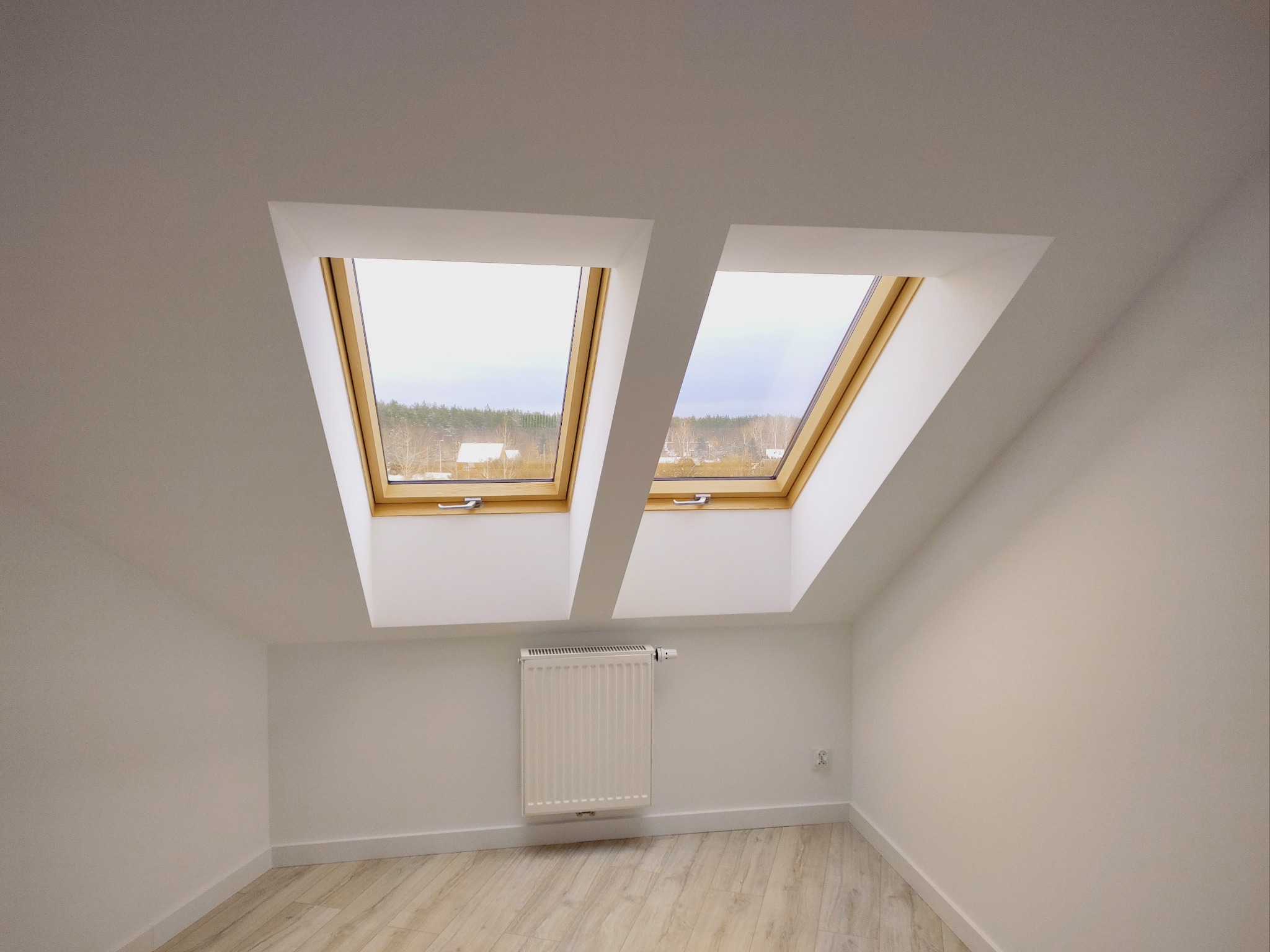Poddasze z dwoma oknami dachowymi z drewnianą ramą, białe ściany, grzejnik i jasna podłoga imitująca drewno, widok na zewnątrz na drzewa.