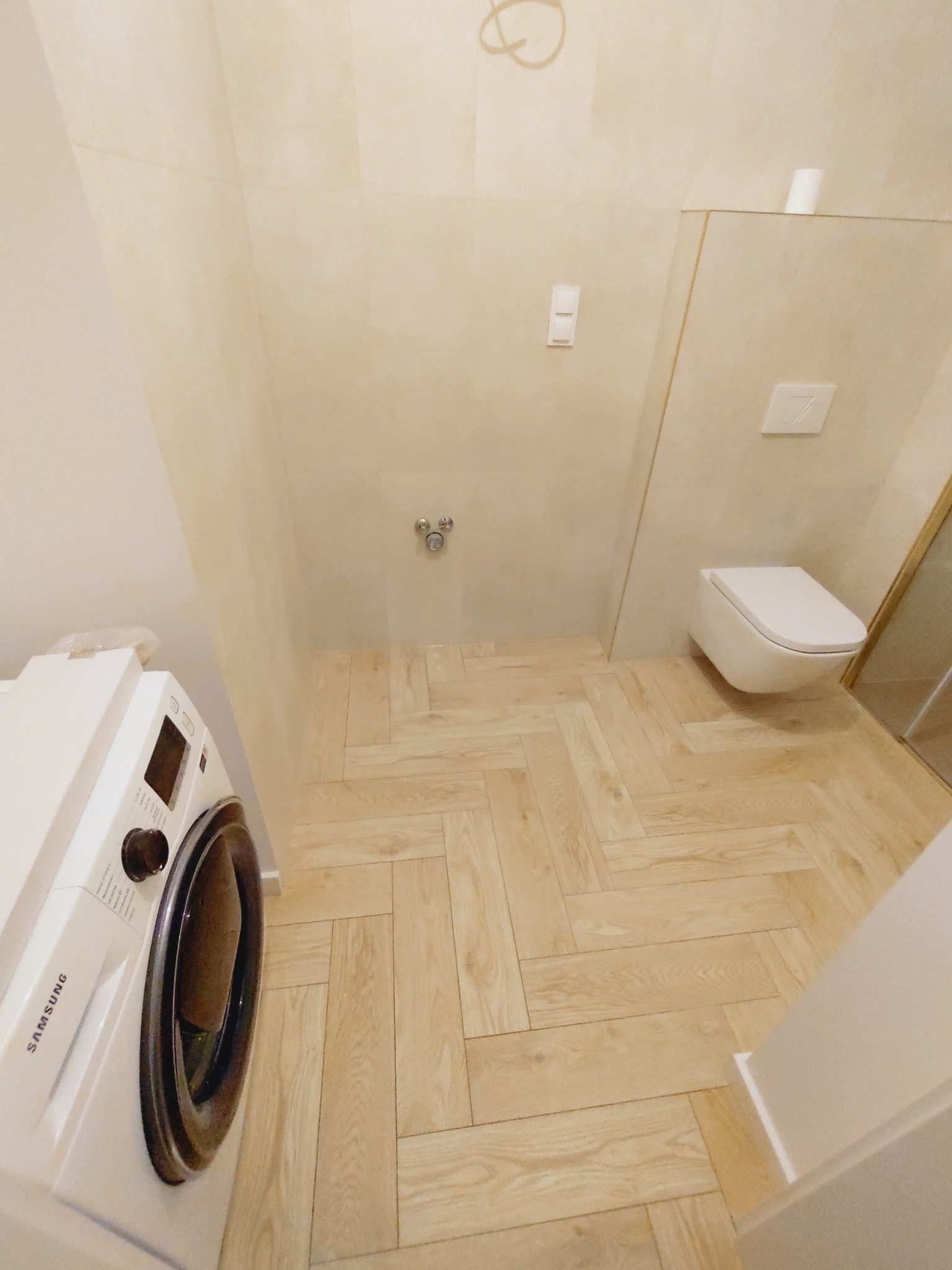 Jasna łazienka z ułożoną w jodełkę podłogą z płytek imitujących drewno, wisząca toaleta i pralka Samsung w rogu.