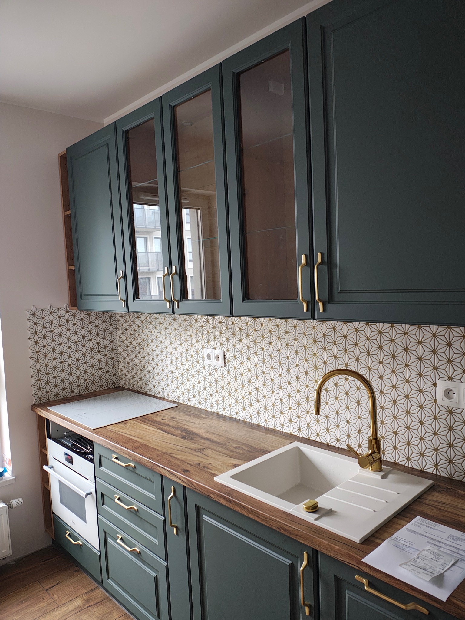 Kuchnia po remoncie: zielone szafki z frezowanymi frontami i złotymi uchwytami, mozaika z białych płytek ze złotą fugą nad blatem z drewnopodobnego laminatu.