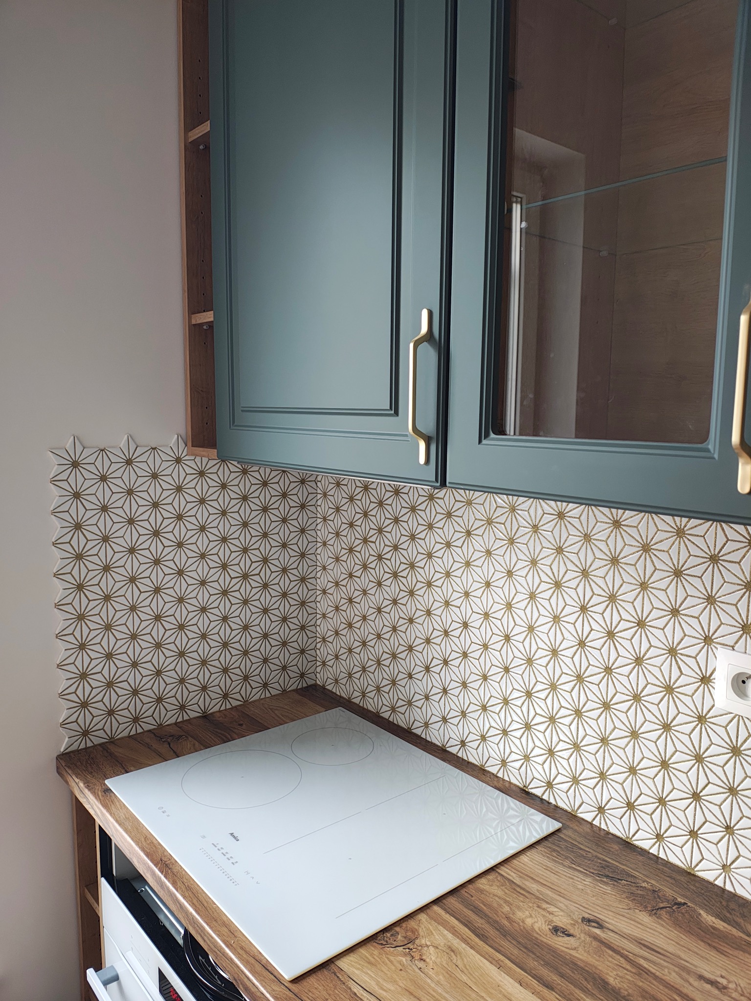 Kuchnia po remoncie: mozaika z geometrycznym wzorem w odcieniach złota i bieli, złote uchwyty szafek, drewniany blat i biała płyta indukcyjna. Nowoczesny design.