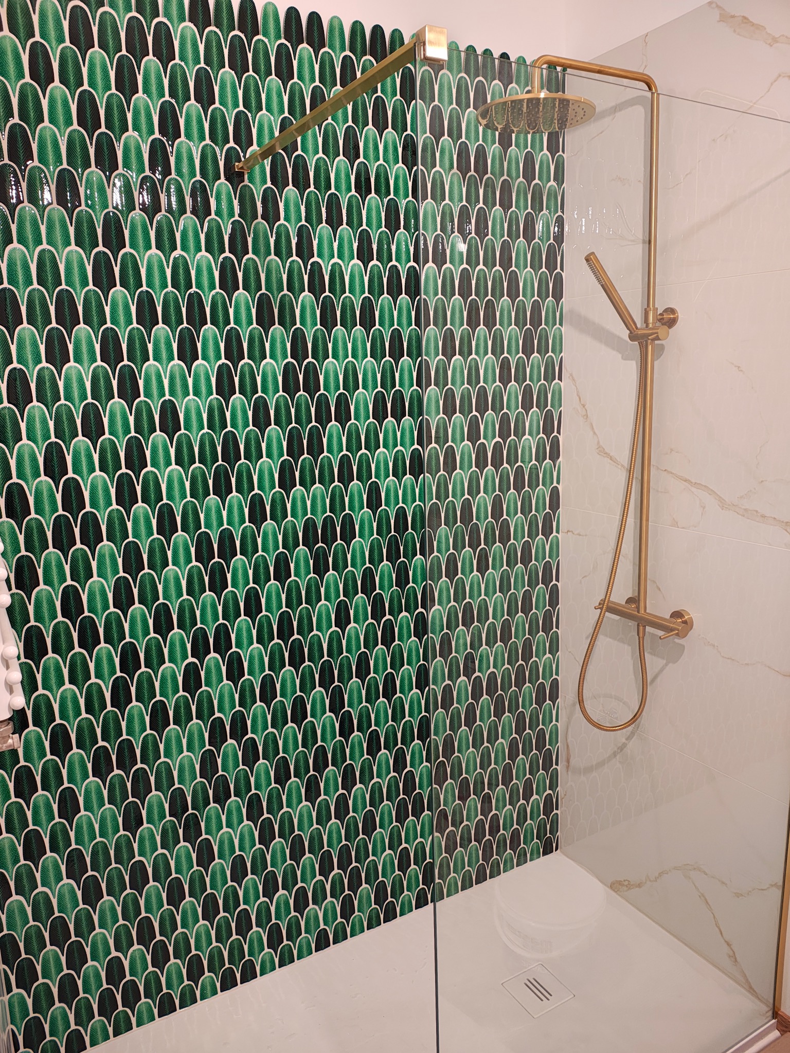Wykończona kabina prysznicowa z zielono-czarną mozaiką w kształcie łusek i złotą armaturą. Biały brodzik z odpływem liniowym. Marmurowe płytki na sąsiedniej ścianie.