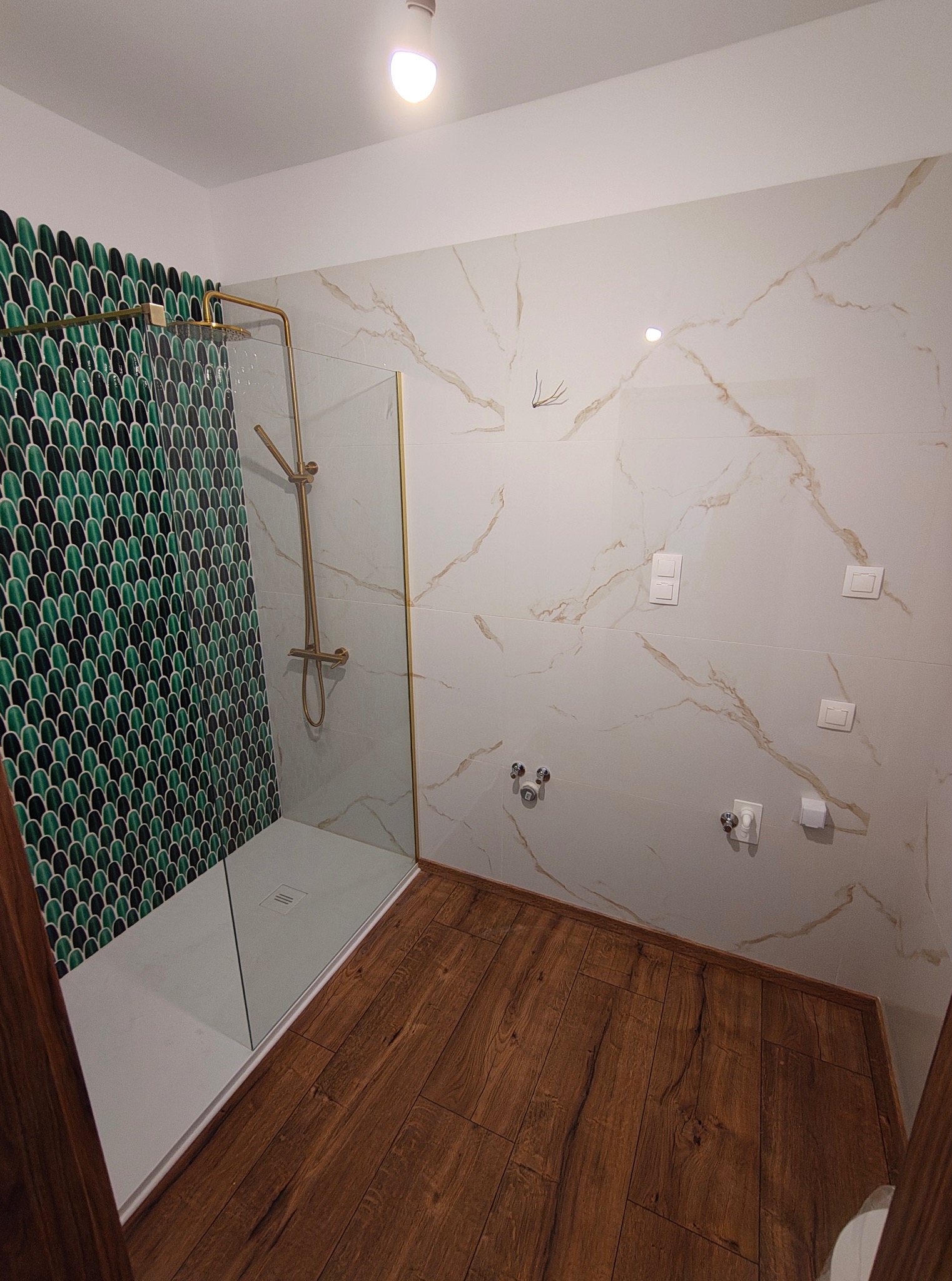 Wykończona łazienka z kabiną prysznicową typu walk-in, zieloną mozaiką w kształcie rybiej łuski, panelami imitującymi marmur i drewnianą podłogą. Złota armatura.