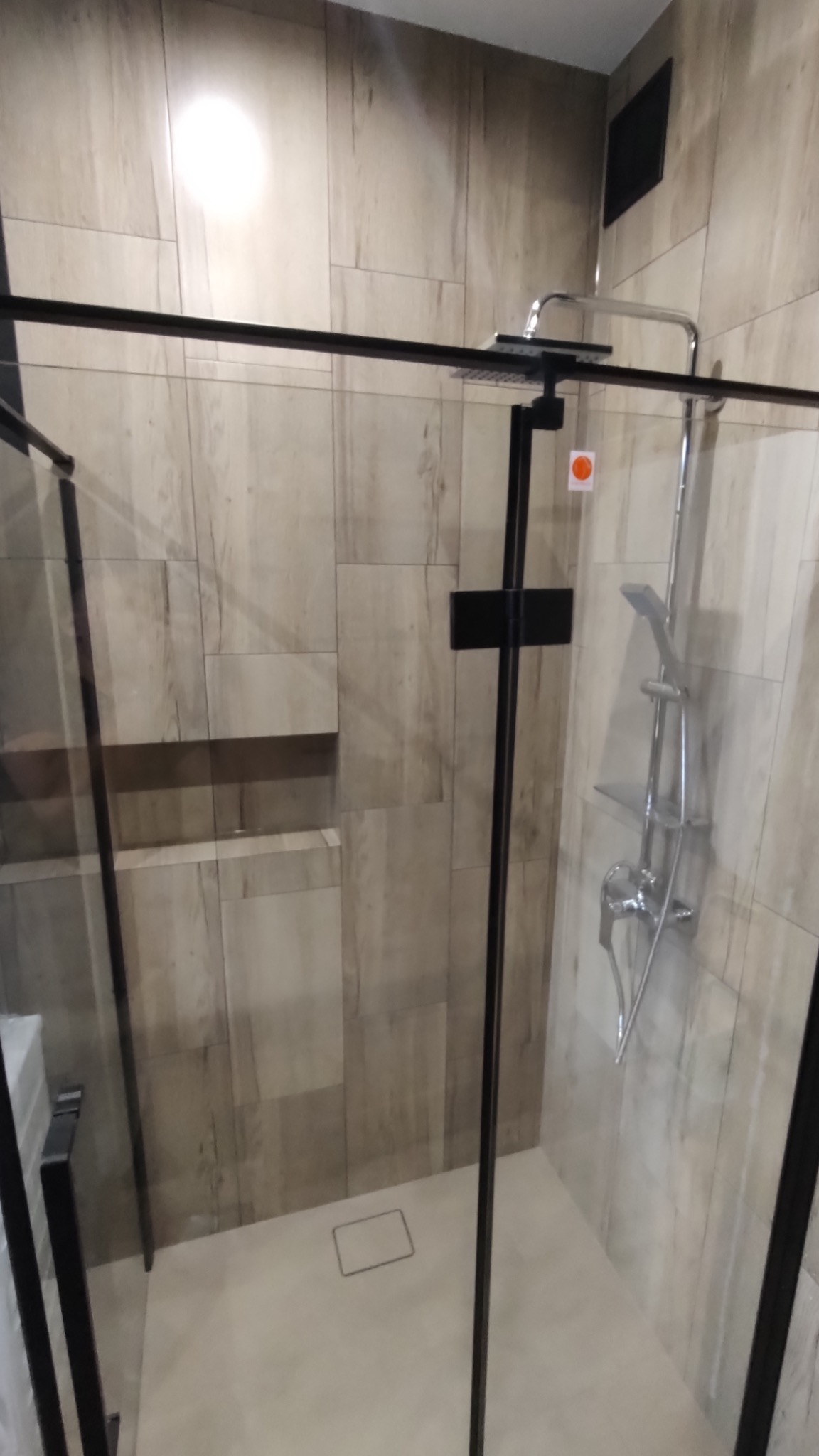 Kabina prysznicowa walk-in z czarnymi profilami, wykończona płytkami imitującymi drewno. Widoczna deszczownica i bateria. Minimalistyczny odpływ liniowy w podłodze.