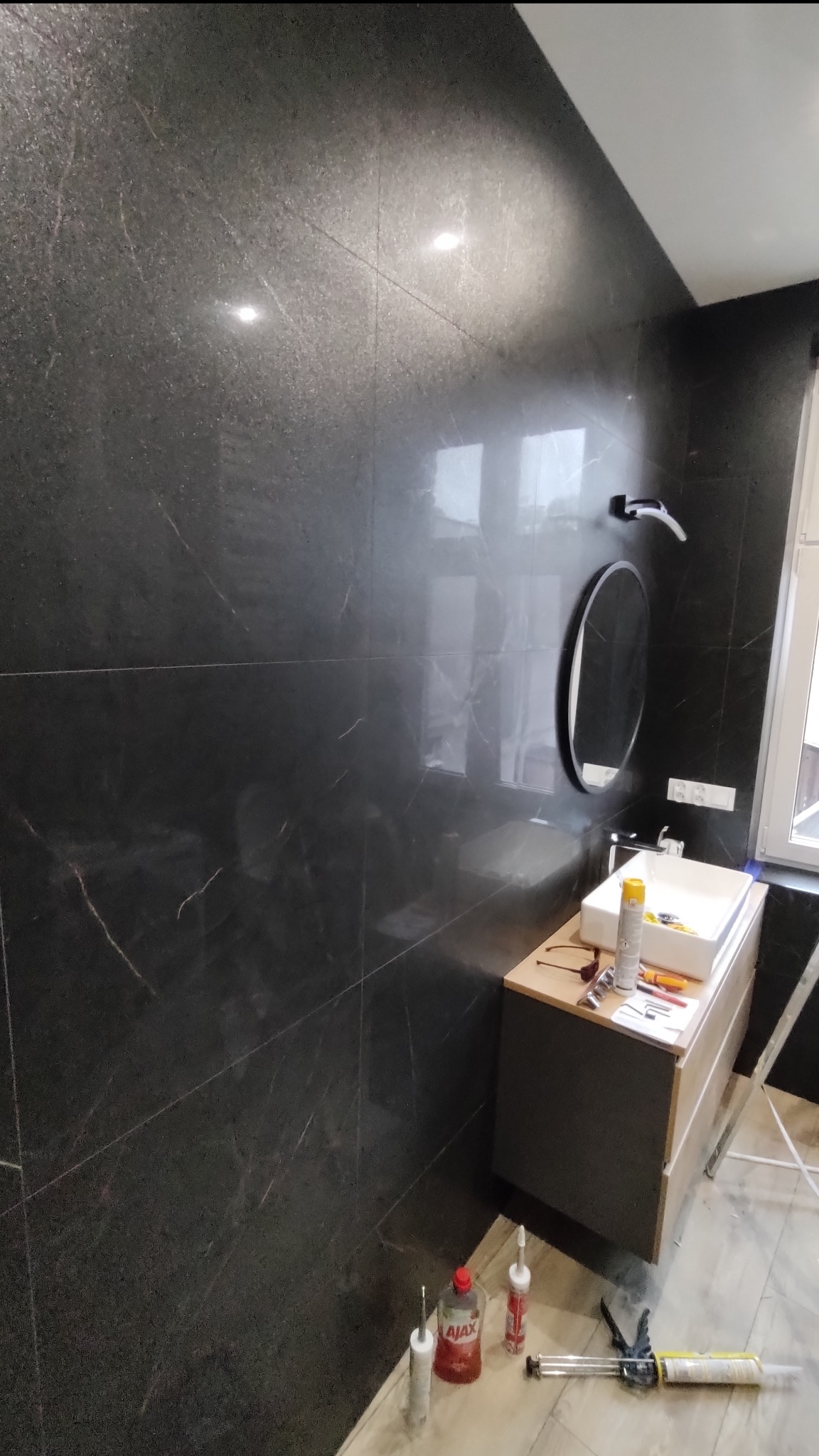 Ciemna, marmurkowa ściana w łazience z okrągłym lustrem i umywalką. Widoczne narzędzia i materiały budowlane, sugerujące etap wykończenia wnętrza.