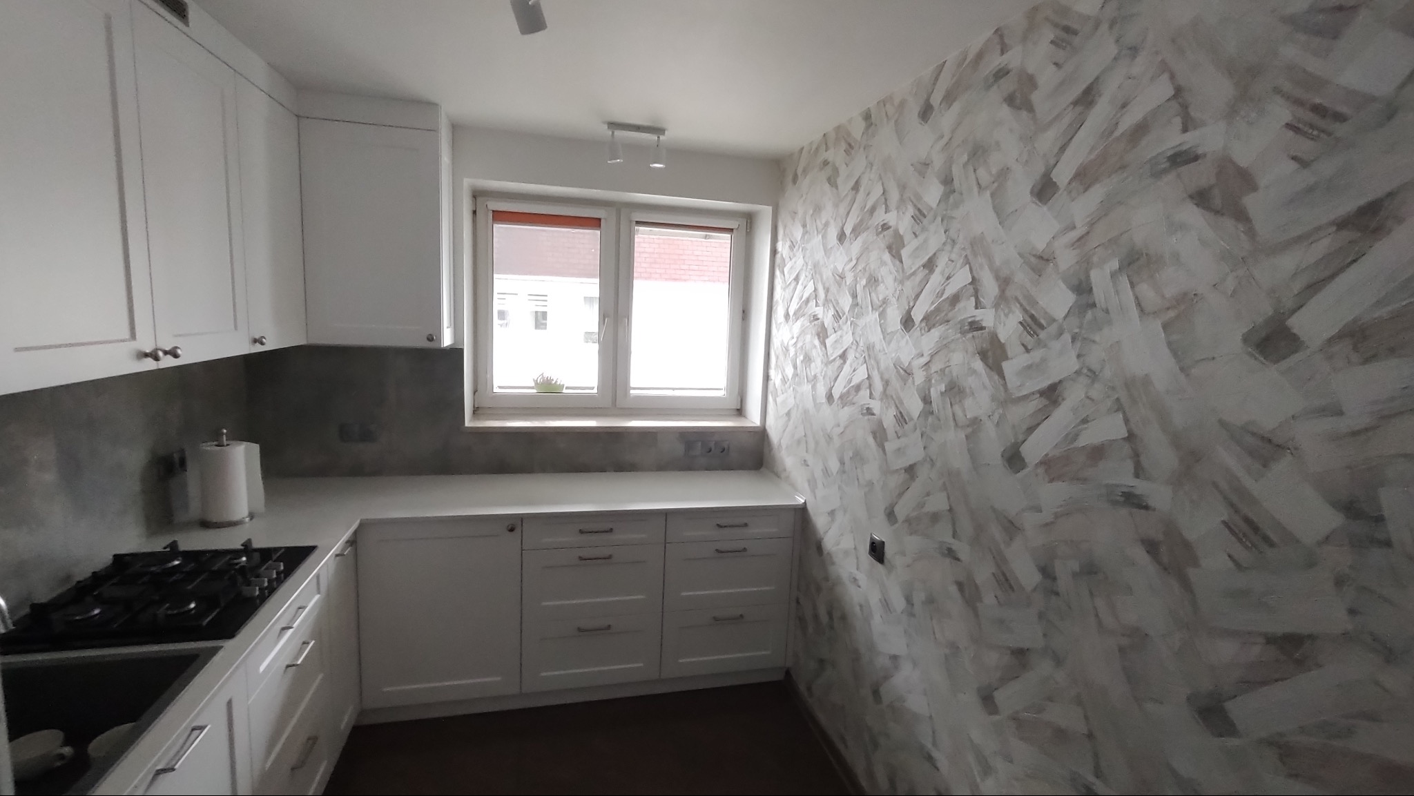 Nowoczesna kuchnia z białymi szafkami i blatem, szarym backsplashiem oraz ścianą z dekoracyjnym malowaniem w odcieniach szarości i bieli. Okno z widokiem na zewnątrz.