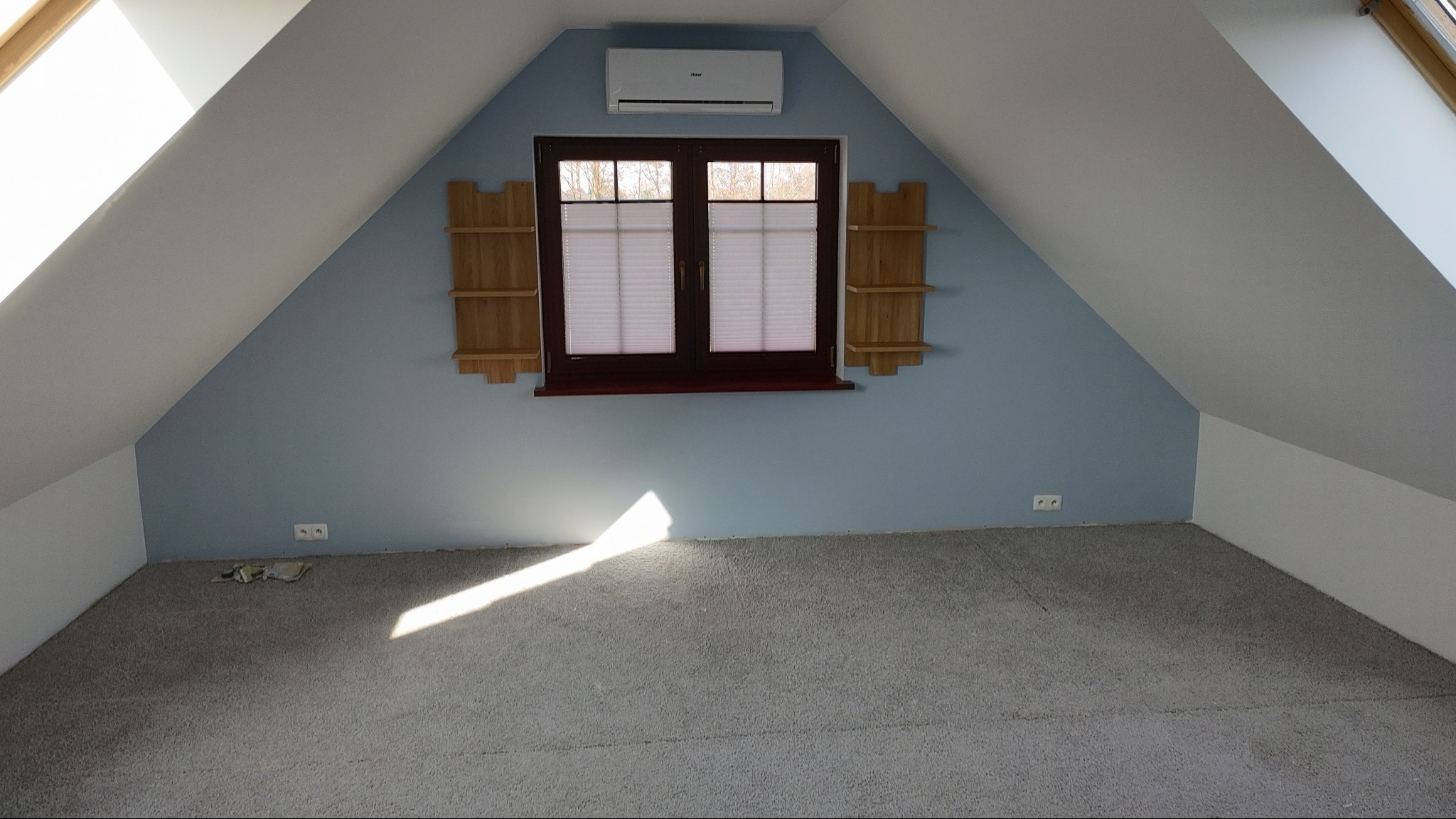 Pokój na poddaszu z oknem, pomalowanymi na niebiesko ścianami i szarą wykładziną. Widoczne drewniane półki po bokach okna i klimatyzator. Świeże malowanie ścian.