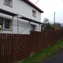 Dwupiętrowy dom z białymi ścianami, zabezpieczonymi oknami i rusztowaniem, widziany zza drewnianego płotu.