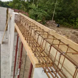 Zbrojenie wieńca ułożone w szalunku z desek na murze z bloczków betonu komórkowego, wsparte stemplami budowlanymi, w tle zieleń drzew.