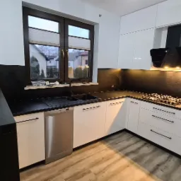 Nowoczesna kuchnia z białymi, lakierowanymi frontami szafek, czarnym blatem i panelem ściennym, z widocznym zlewem, płytą grzewczą i okapem, oświetlona ciepłym światłem.