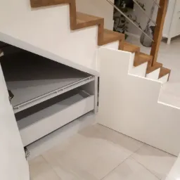 Białe schody z drewnianymi stopniami i wbudowaną szafką z szufladami, otwartą po lewej stronie, ukazując wnętrze z szufladami.