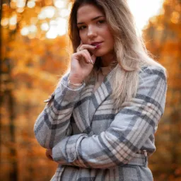 Portret młodej kobiety w jesiennej scenerii, ubrana w szary płaszcz w kratę, z dłonią przy ustach i złotym pierścionkiem na palcu, z rozmytym pomarańczowym tłem.