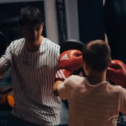 Dwóch chłopców trenuje boks w pomieszczeniu: jeden trzyma czarne tarcze, drugi uderza w nie czerwonymi rękawicami Adidas.