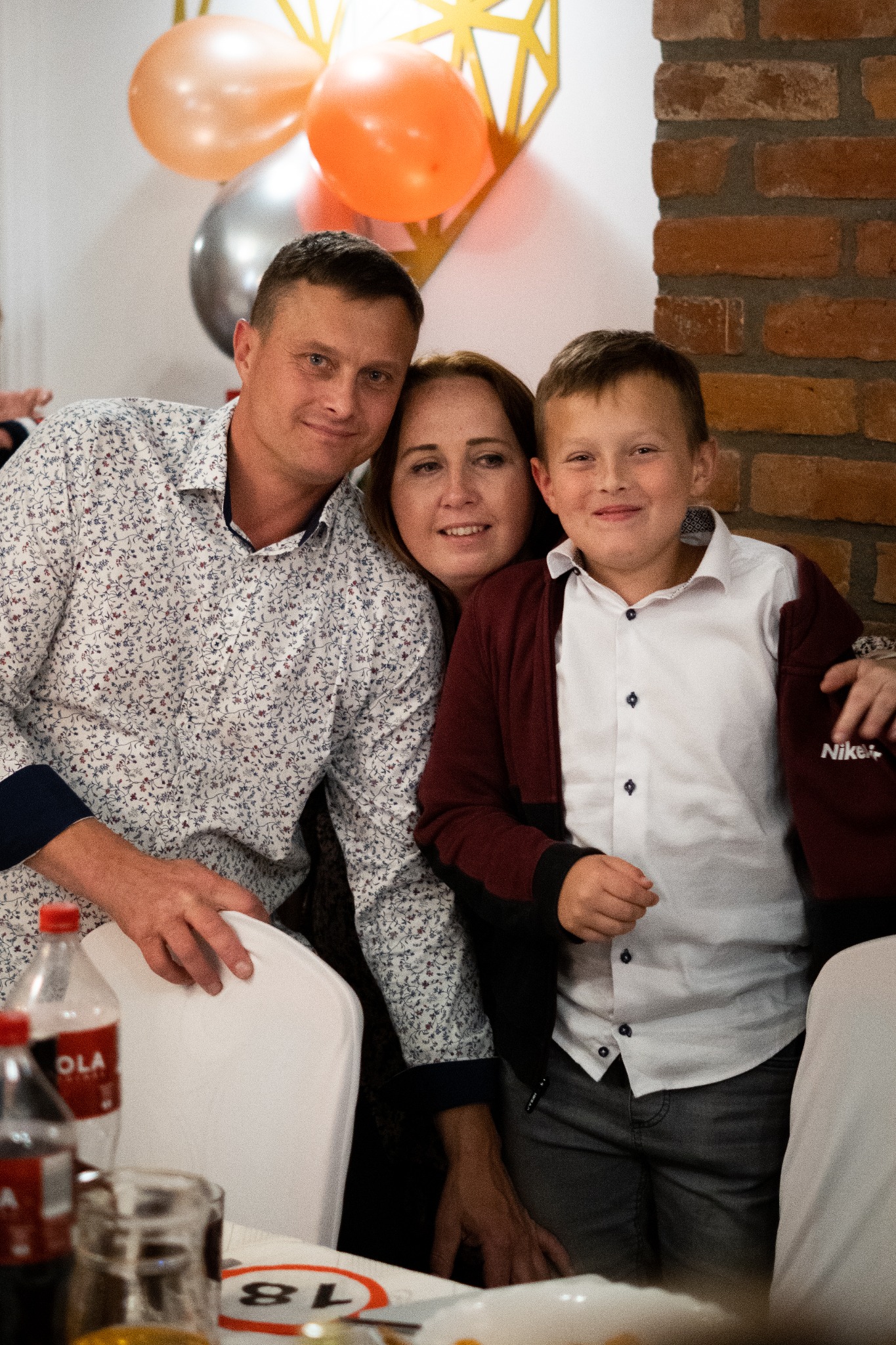 Uśmiechnięta rodzina: ojciec w kwiecistej koszuli, matka i syn w bordowej bluzie na tle ceglanej ściany i dekoracji balonowych podczas uroczystości.