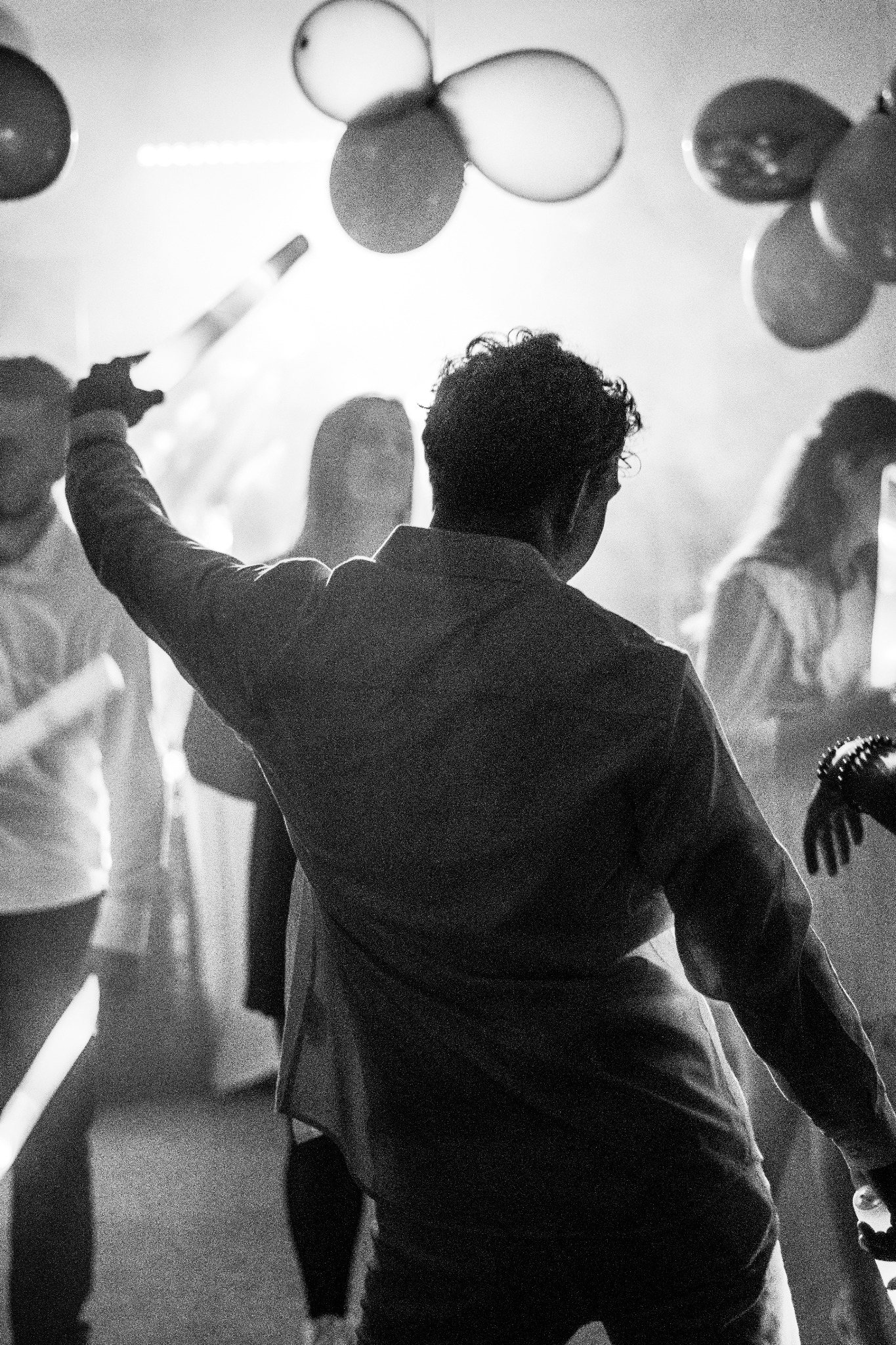 Czarno-białe zdjęcie z imprezy, mężczyzna tyłem z uniesioną ręką trzymający świecący przedmiot, w tle balony i zamazane sylwetki gości.