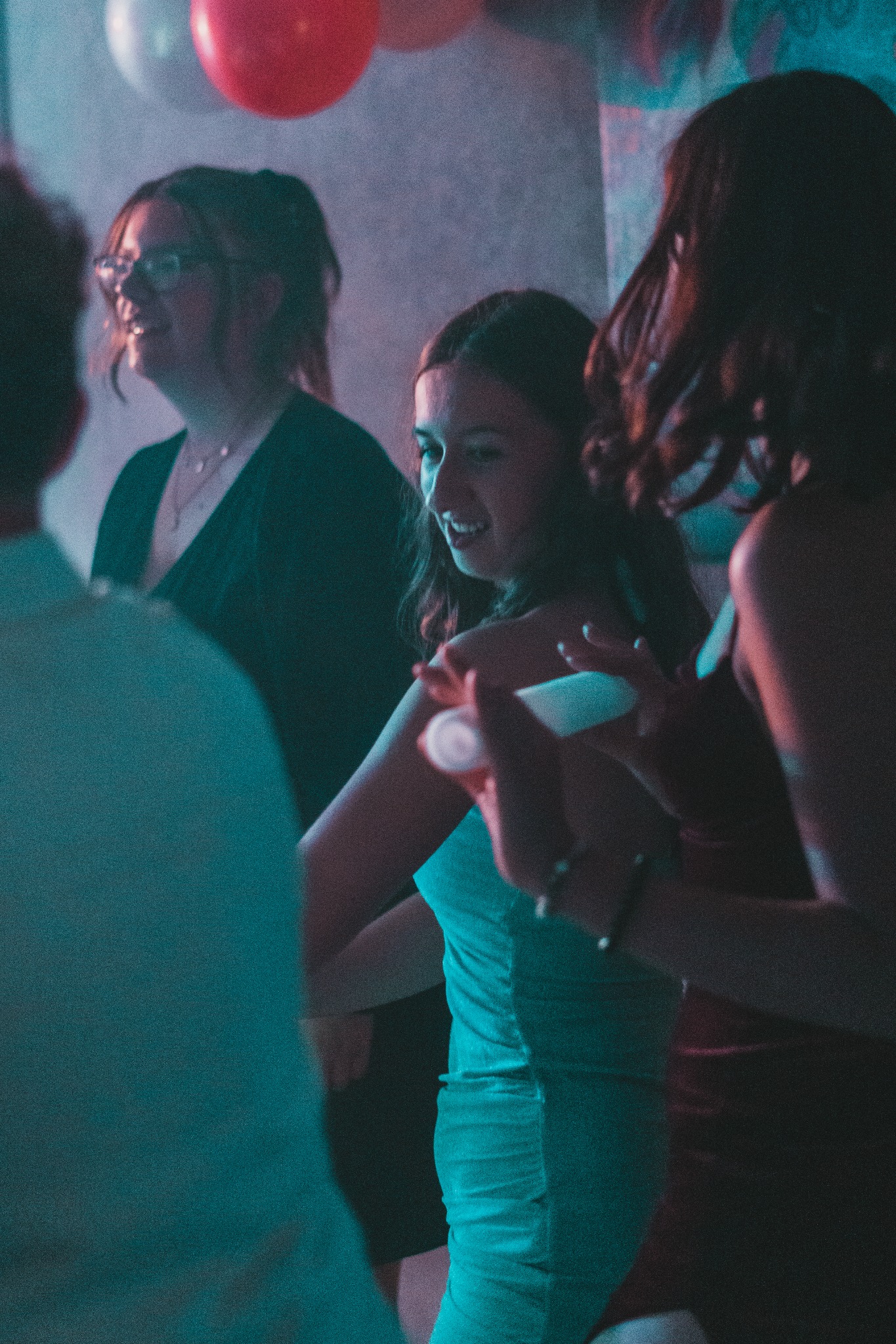 Młoda kobieta w turkusowej sukience uśmiecha się podczas imprezy, trzymając mały, biały przedmiot w dłoni, w tle widoczne balony i inne osoby.