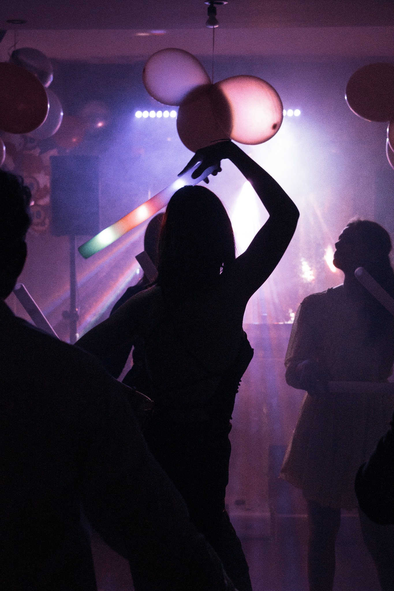 Silhouettes of dancing people with glowing light sticks and balloons at a party with purple lighting and a DJ in the background.