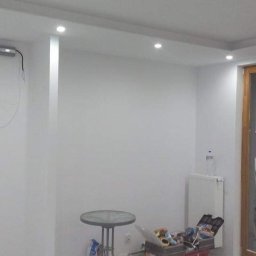 ROMBUD - Pokój po odświeżeniu ścian w jasnych barwach, widoczne nowe oświetlenie sufitowe, okno balkonowe z widokiem na budynek, mały stolik, grzejnik i skrzynka z narzędziami w rogu.