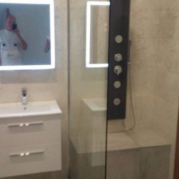 ROMBUD - Nowoczesna łazienka z kabiną prysznicową typu walk-in z czarnym panelem natryskowym, umywalką z szafką i lustrami z podświetleniem LED, widoczne odbicie osoby robiącej zdjęcie w lustrze.