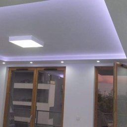 ROMBUD - Nowoczesny salon z podwieszanym sufitem, oświetleniem LED i drewnianymi drzwiami balkonowymi z widokiem na miasto.