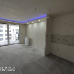 ROMBUD - Surowe wnętrze mieszkania w stanie deweloperskim z wylewką, grzejnikiem żeberkowym, grzejnikiem płytowym, marmurową mozaiką na ścianie i podwieszanym sufitem z niebieskim oświetleniem LED.