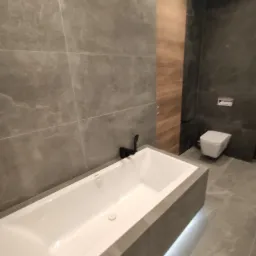 Nowoczesna łazienka w Warszawie z wanną obudowaną szarymi płytkami, czarną armaturą i podświetleniem LED pod wanną, widok na wiszącą toaletę.